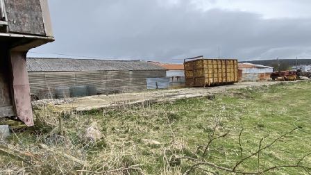 A photograph showing a rural site featuring a long corrugated metal agricultural building and a large skip in the foreground.