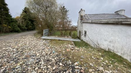 A photograph showing a white-walled building with a slate roof on the right, a gravel driveway in the foreground, and a metal gate leading to a grassy area.