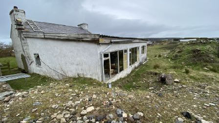 A photograph of an existing single-story white dwelling with a large windowed extension and solar panels on the roof, set in a rural landscape.