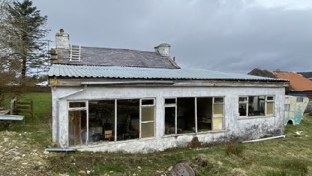 A photograph of an existing single-story white bungalow with a slate roof, appearing to be in a state of disrepair or renovation with a ladder leaning against the chimney stack.