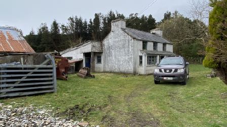 A photograph showing a rural property with an existing white cottage-style dwelling and a corrugated metal outbuilding. A dark SUV is parked on the grass in front of the house near a metal gate.