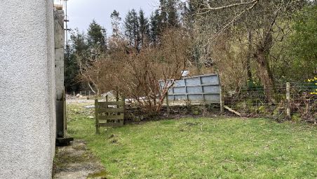A photograph showing the rear garden area of a property with a white wall on the left, a wooden fence, and trees in the background.