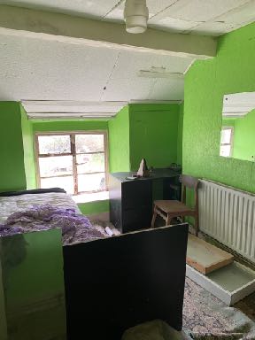 Interior photograph of a room with bright green walls, showing a bed, desk, and window looking out to a garden.
