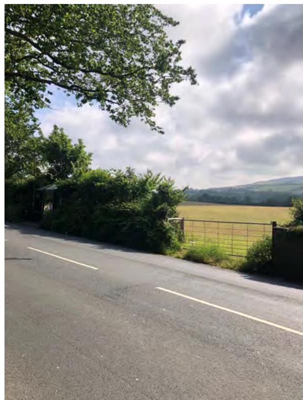 A photograph showing a rural road with a bus shelter on the left and a field gate on the right, depicting the existing site conditions.