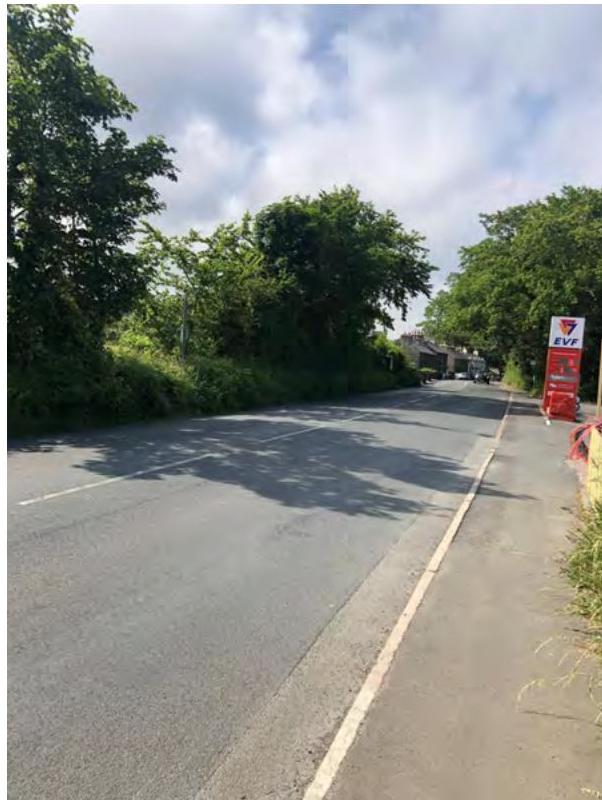 A photograph showing a paved rural road with dense trees and vegetation on both sides and a red sign on the right.