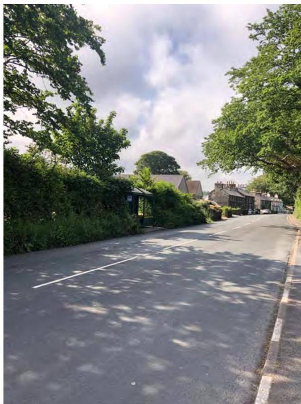 A street-level photograph showing a bus shelter on the side of a rural road with houses and trees in the background.