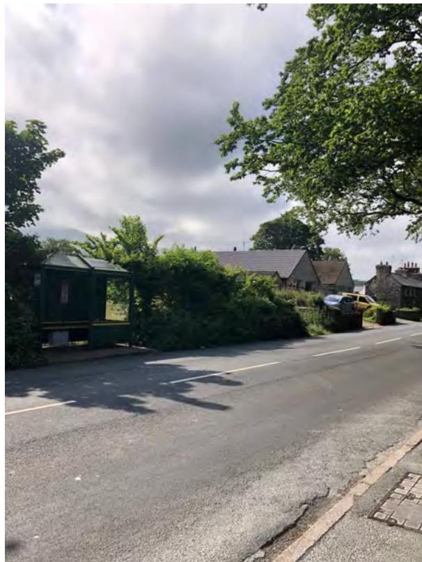 A street-level photograph showing a green bus shelter on the left side of a road, with houses and trees in the background.