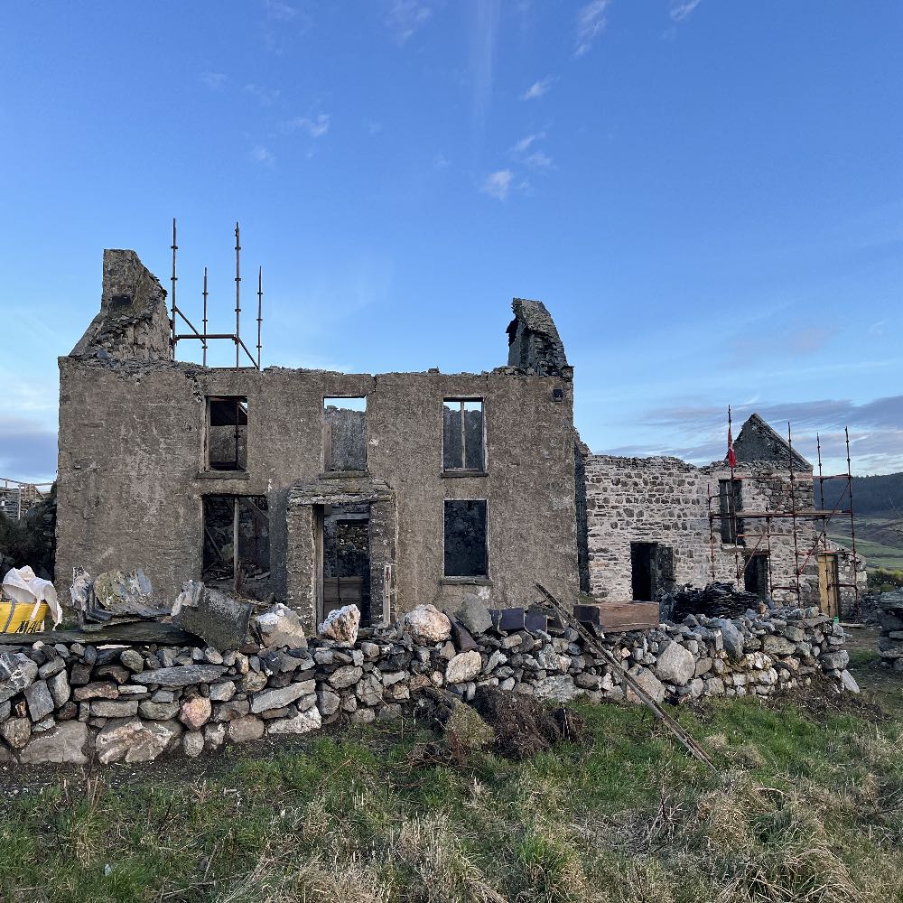 A photograph showing a derelict stone building with scaffolding and exposed window openings, located behind a dry stone wall in a rural landscape.