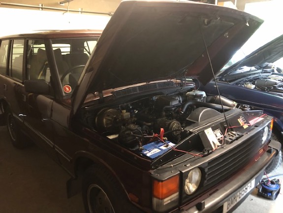 A photograph of a maroon car with its hood open inside a garage, showing the engine and a battery charger.