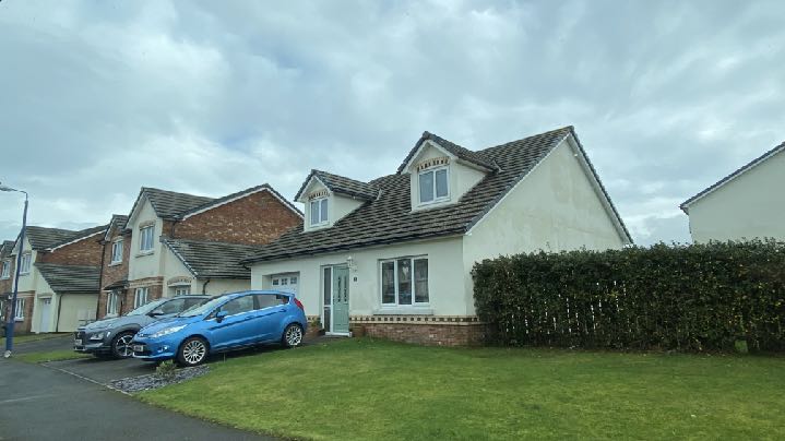 A street-level photograph showing a white detached house with dormer windows and a tiled roof, situated next to brick semi-detached houses with cars parked in front.