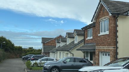 A street-level photograph showing a row of modern brick and rendered semi-detached houses with cars parked in front under a blue sky.