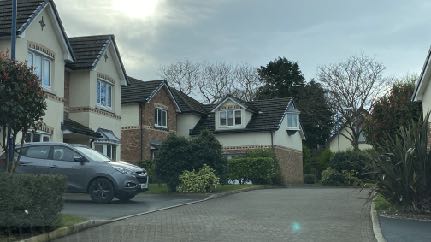 A street-level photograph showing a row of residential houses with driveways, parked cars, and surrounding greenery.