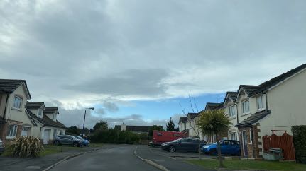 A street-level photograph showing a residential road with houses on either side and several parked cars under a cloudy sky.