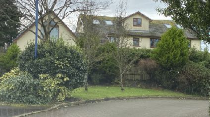 A photograph showing a detached residential property with a pitched roof and dormer windows, surrounded by mature trees and shrubbery.