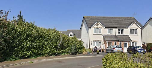 A street-level photograph showing a white two-story house with a driveway and parked cars, bordered by a large green hedge on the left.