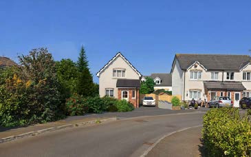 A street-level photograph showing existing detached houses and parked cars on a residential road under a clear blue sky.