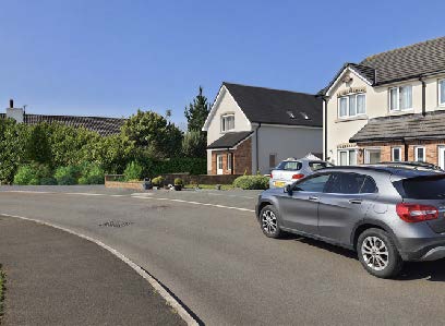 A street-level photograph showing a grey car parked on a road in front of residential properties, including a white-rendered detached house. The view captures the existing site context and surrounding road infrastruct...