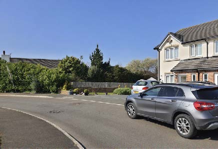 A street-level photograph showing a residential road with a parked car, a two-story house on the right, and a wooden fence with vegetation behind it.