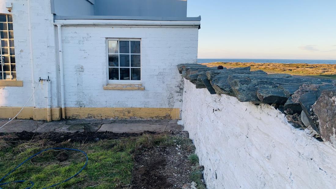 Exterior photograph of a white painted building with a window, situated next to a stone wall with the sea visible in the background.