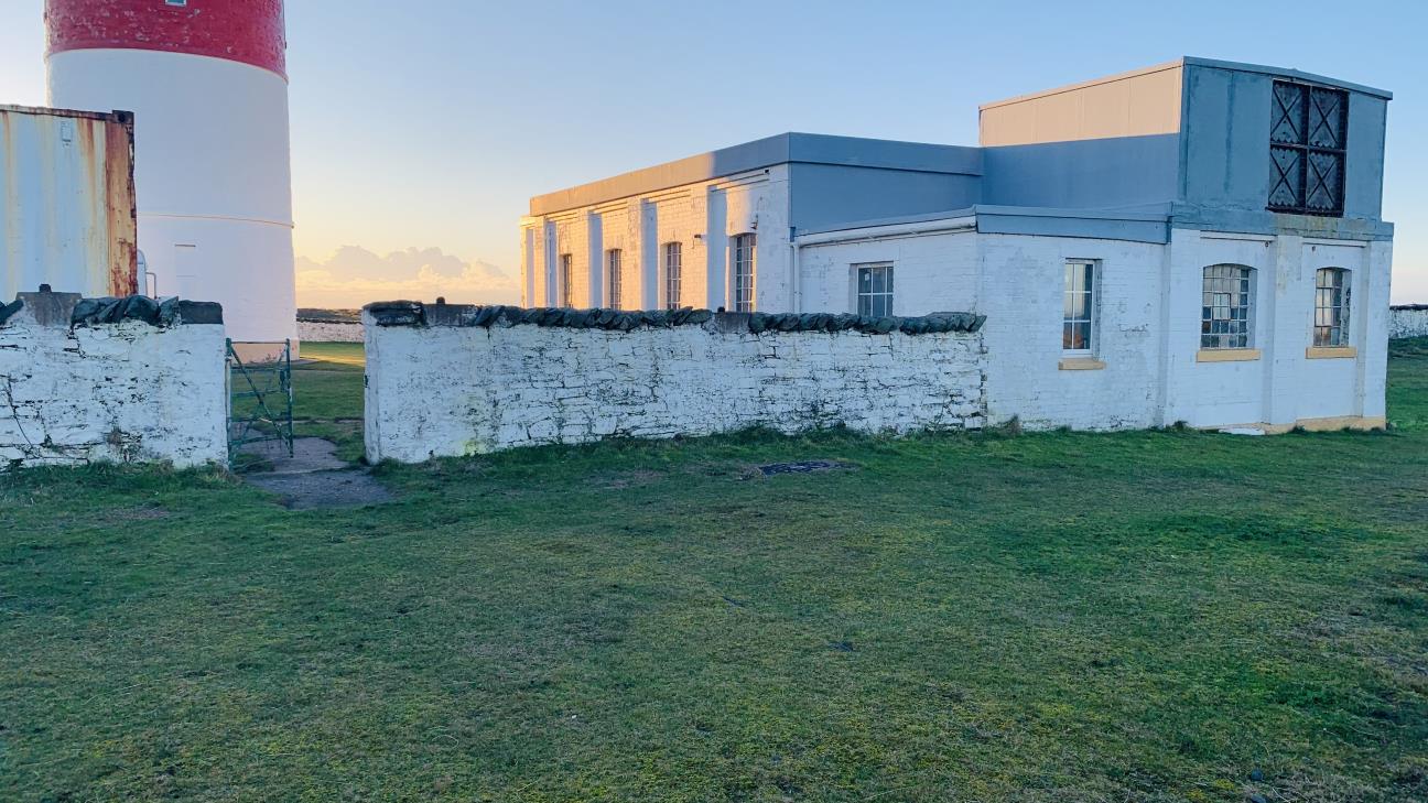A photograph showing a white outbuilding with a grey roof extension situated next to a red and white lighthouse tower. A white stone wall runs along the foreground with grassy ground.