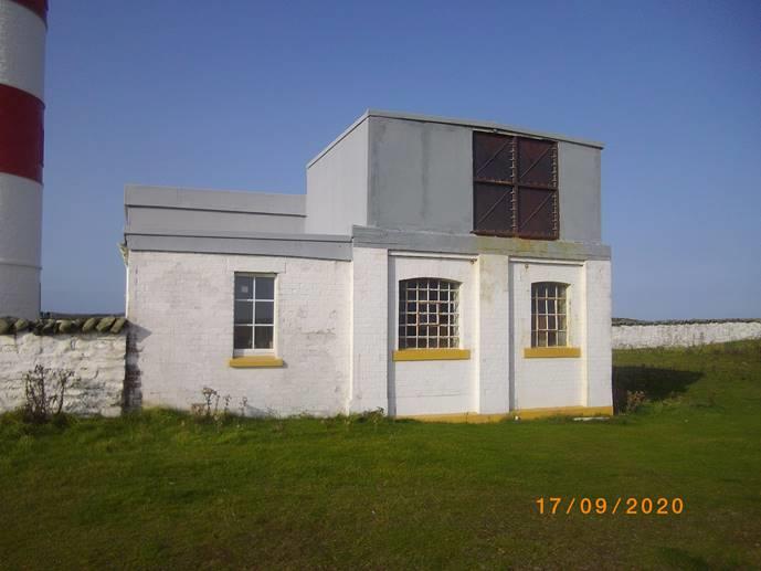 A photograph showing a white single-story outbuilding with a grey upper section located next to a red and white lighthouse tower.