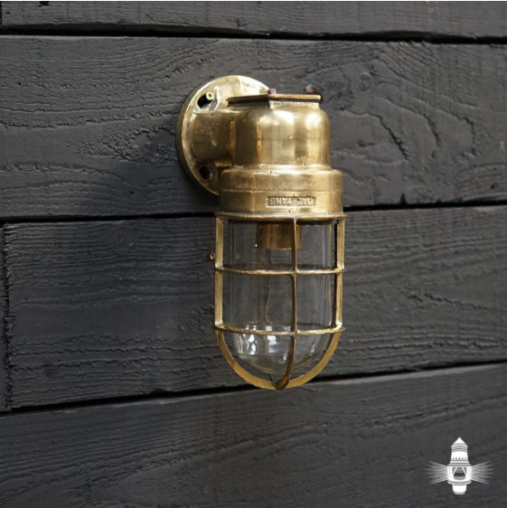 A close-up photograph of a brass outdoor security light fixture mounted on dark, weathered wooden cladding.