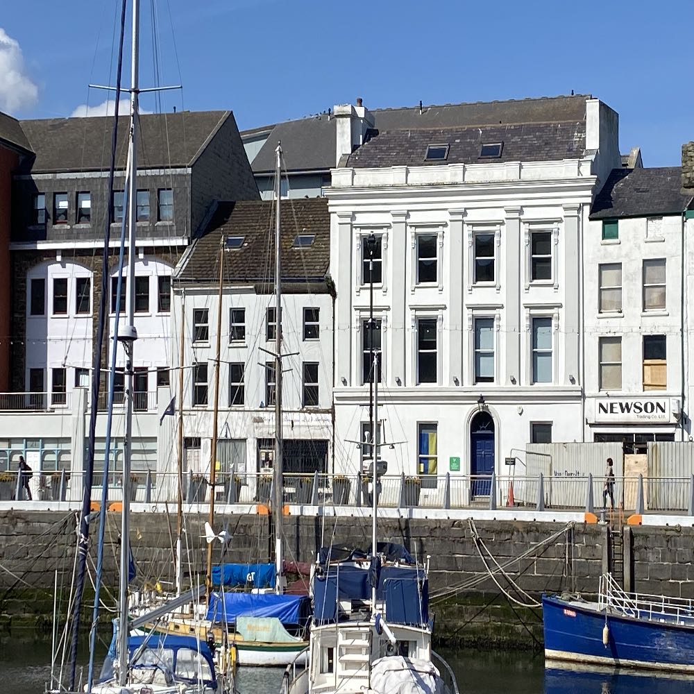 A photograph showing a row of buildings along a harbor quay with boats in the foreground, featuring a prominent white commercial building with 'NEWSON' signage.