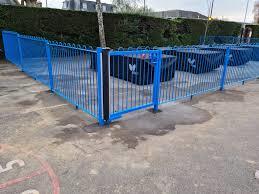 A photograph showing a blue metal fence enclosing a skate park area with concrete ramps and a hedge in the background.