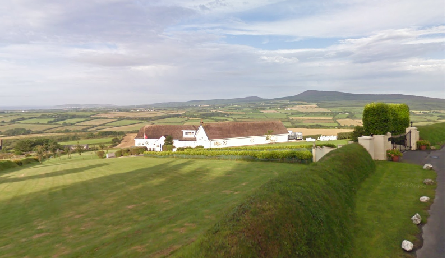 A high-angle photograph showing a white detached bungalow situated on a grassy slope within a rural landscape.