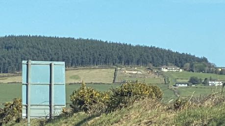 A photograph of a rural landscape featuring a large coniferous forest on a hill in the background. The foreground shows a grassy verge with a blank sign board and scattered houses in the distance.