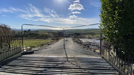 A photograph taken from a driveway looking out through an open metal gate towards a rural landscape with coastal views in the distance.