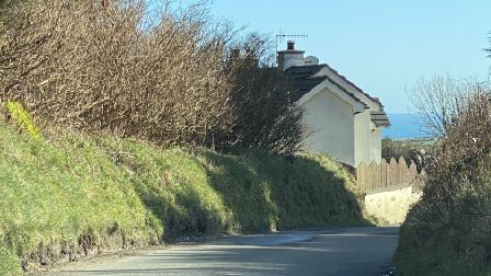 A street-level photograph showing a white single-story bungalow situated on a grassy embankment next to a road, with a view of the sea in the background.