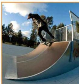 A photograph showing a skateboarder performing a trick on a curved ramp at a skate park, with a metal fence and trees in the background.