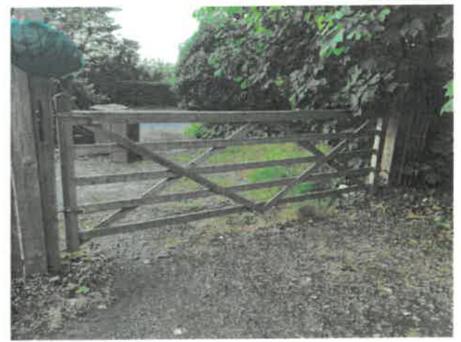 A photograph showing a wooden field gate at the entrance of a property with a gravel driveway and surrounding vegetation.