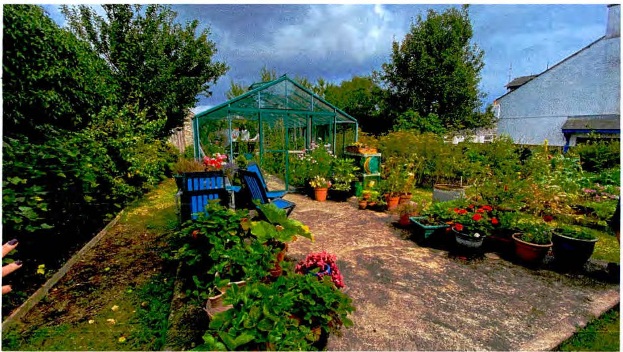 A photograph showing a residential garden featuring a large greenhouse structure, numerous potted plants, and a paved patio area.