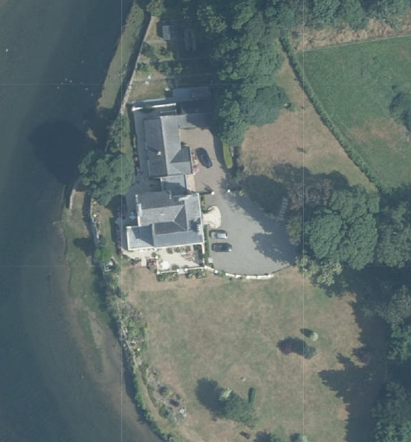 An aerial view of a detached house situated on a coastal plot, showing the main building, driveway with vehicles, and surrounding fields.
