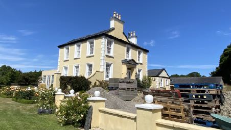 A photograph showing a large, cream-colored detached house with a garden and boundary wall in the foreground, with stacked materials visible to the right.