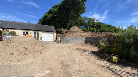 A photograph showing a rural property with a bungalow and an existing garage on the left. The foreground features a large pile of sand and a low stone wall under construction.