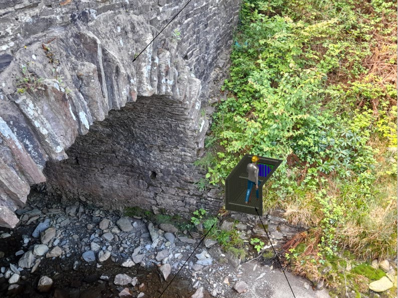 A site photograph of a stone arch bridge or culvert over a rocky stream bed, featuring a digital 3D overlay illustrating the proposed energy dissipater installation.