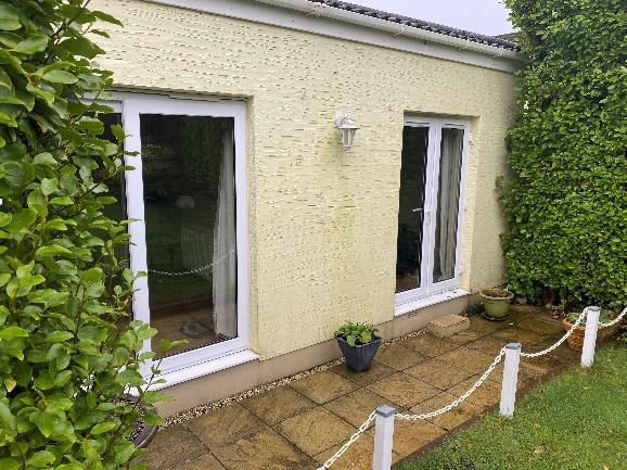 A photograph showing the exterior rear elevation of a single-story building with cream rendered walls, white-framed windows, and a paved patio area bordered by hedges.