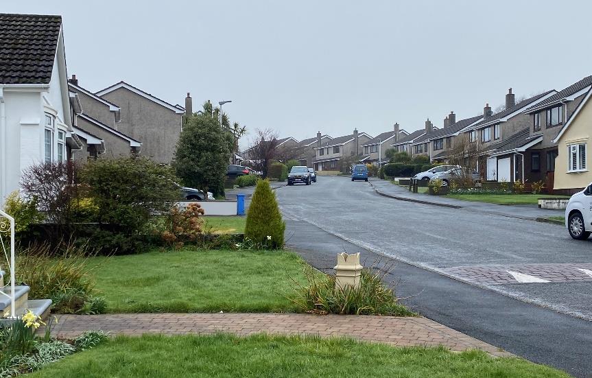 A street-level photograph showing a residential road lined with semi-detached houses and parked cars. The view is taken from a front garden looking down the street towards the distance.