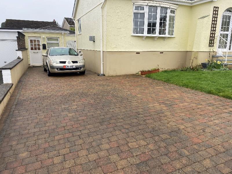A photograph showing a brick-paved driveway with a parked car, adjacent to a cream-colored house and a white garage or outbuilding structure.