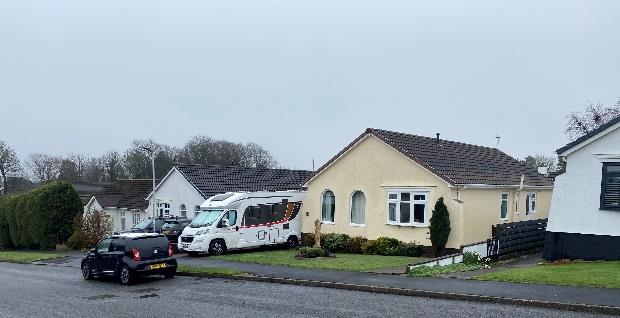 A street-level photograph showing a cream-colored detached bungalow with a large caravan parked in the front driveway and a car on the road.