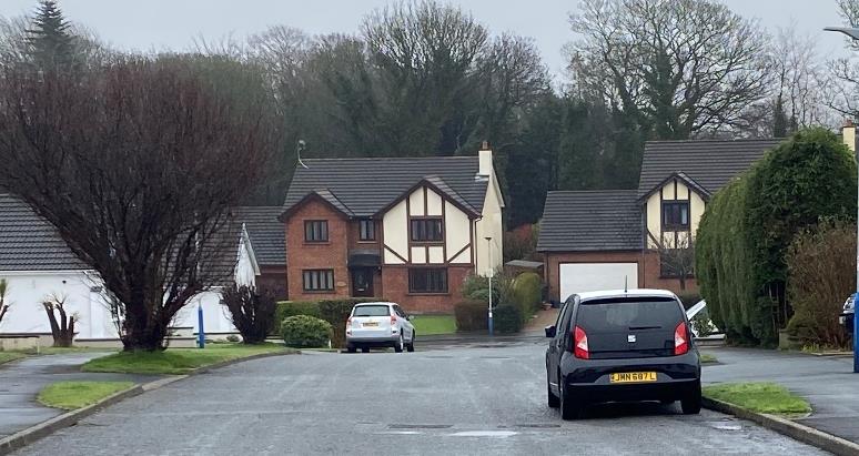A street-level photograph showing a row of detached houses with brick and timber-framed facades. Cars are parked on the wet road in front of the properties, surrounded by trees and hedges.