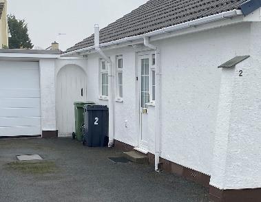 A photograph showing the side elevation and driveway of a white single-story bungalow. The image features an attached garage, a main entrance door, windows, and two wheelie bins on the paved forecourt.