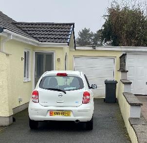 A photograph showing the side elevation of a cream-colored bungalow with a driveway, a parked white car, and an attached garage with a white door.