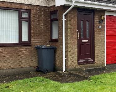 A close-up photograph of a brick residential property exterior featuring a brown front door, windows with dark frames, and a bright red garage door.