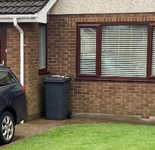 A photograph showing the brick exterior wall of a residential property, featuring a window, a wheelie bin, and a parked car.