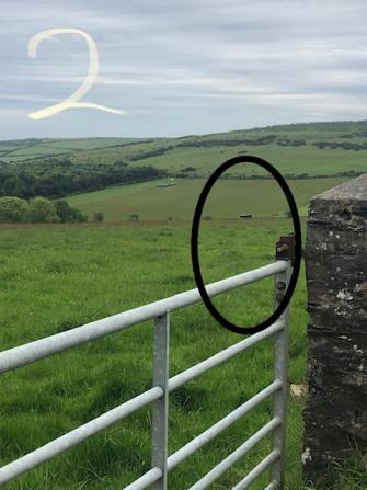 A photograph of a rural green field with a metal gate and stone wall in the foreground. A black circle highlights a specific area in the distance, likely indicating the proposed site or existing feature.
