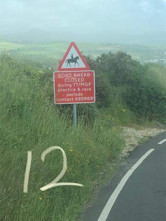 A photograph showing a red road sign warning of a horse rider ahead and road closures for TT/MGP events, set against a rural hillside with a large number 12 written in the grass.
